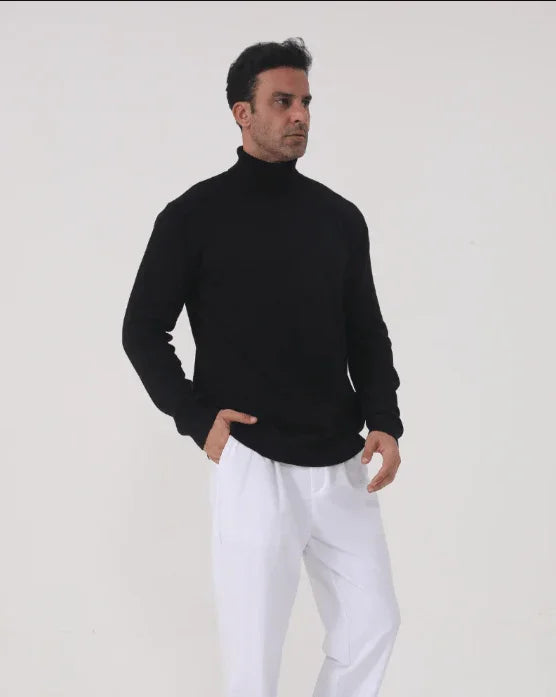 Man in black turtleneck sweater and white pants posing against plain white background
